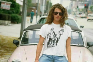 A man with long curly hair and sunglasses stands in front of a vintage convertible car on a city street, wearing a white T-shirt with a black graphic print and blue jeans.