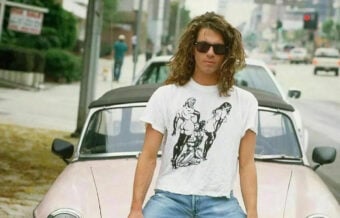 A man with long curly hair and sunglasses stands in front of a vintage convertible car on a city street, wearing a white T-shirt with a black graphic print and blue jeans.