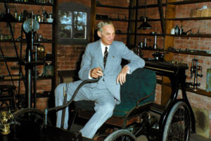 A man in a light gray suit sits in an old-fashioned car inside a workshop filled with shelves, laboratory equipment, and tools, with brick walls and a window in the background.
