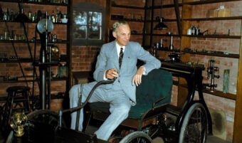 A man in a light gray suit sits in an old-fashioned car inside a workshop filled with shelves, laboratory equipment, and tools, with brick walls and a window in the background.