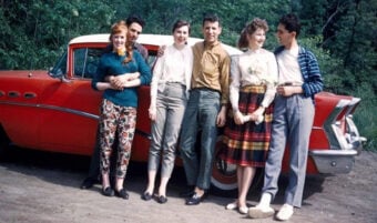 Six young adults stand smiling in front of a vintage red car, dressed in 1950s-style clothing, with trees and greenery in the background.