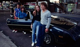 Four young people pose on a city street next to a black sports car with an ornate gold design on the hood. Two are leaning against the car, and two are standing close together, smiling.