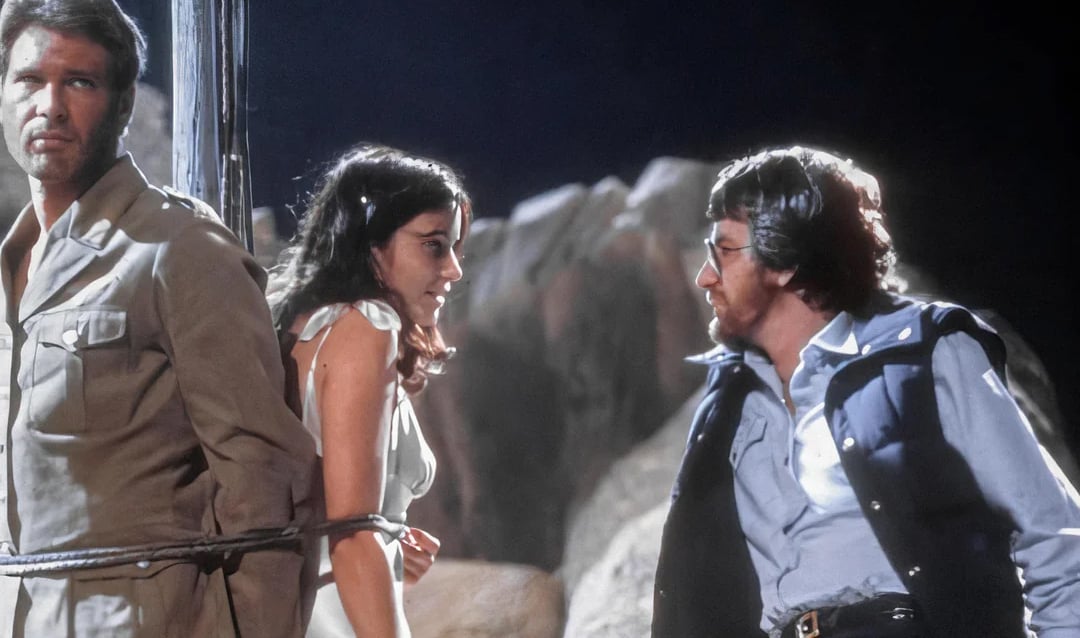 A man and woman are tied together with rope while another man faces them, speaking seriously. They are outdoors, surrounded by large rocks, and it appears to be nighttime.