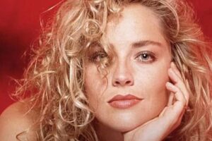 A woman with curly blonde hair rests her face on her hand, gazing at the camera with a relaxed expression. The background is a deep red, complementing her lipstick and soft makeup.