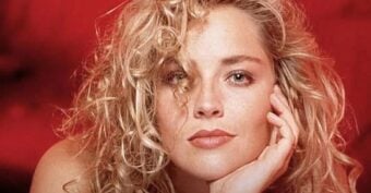 A woman with curly blonde hair rests her face on her hand, gazing at the camera with a relaxed expression. The background is a deep red, complementing her lipstick and soft makeup.