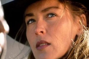 A close-up of a woman with light brown hair wearing a wide-brimmed black hat, looking thoughtfully to the side. Another blurred figure is partially visible in the foreground.