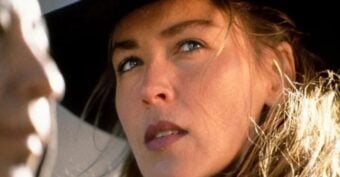 A close-up of a woman with light brown hair wearing a wide-brimmed black hat, looking thoughtfully to the side. Another blurred figure is partially visible in the foreground.