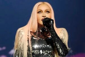 A performer with long, straight, pink-blond hair sings into a microphone while wearing a black, shiny, geometric-patterned outfit against a dark, moody background.