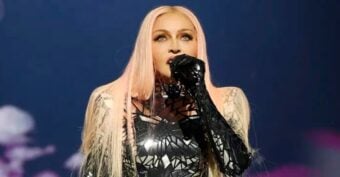 A performer with long, straight, pink-blond hair sings into a microphone while wearing a black, shiny, geometric-patterned outfit against a dark, moody background.