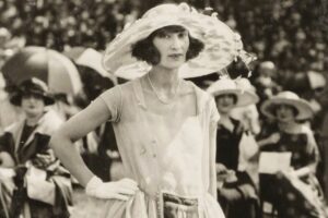 A woman in a light dress, gloves, and a large hat stands confidently outdoors at a formal event, with a crowd of similarly dressed people seated and wearing hats in the background. The photo is in black and white.