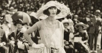 A woman in a light dress, gloves, and a large hat stands confidently outdoors at a formal event, with a crowd of similarly dressed people seated and wearing hats in the background. The photo is in black and white.
