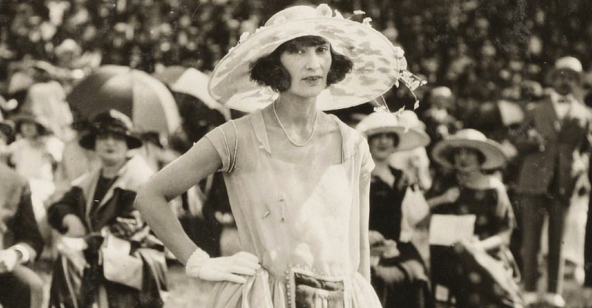 A woman in a light dress, gloves, and a large hat stands confidently outdoors at a formal event, with a crowd of similarly dressed people seated and wearing hats in the background. The photo is in black and white.