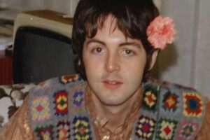 A young man with dark hair wears a colorful crocheted vest and has a pink flower tucked behind his ear. He sits indoors on a patterned sofa, looking directly at the camera.