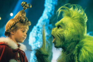 A young girl in a red and white coat faces a green, furry creature with a surprised expression in a festive, whimsical setting. The girl has an elaborate hairstyle decorated with a bow.