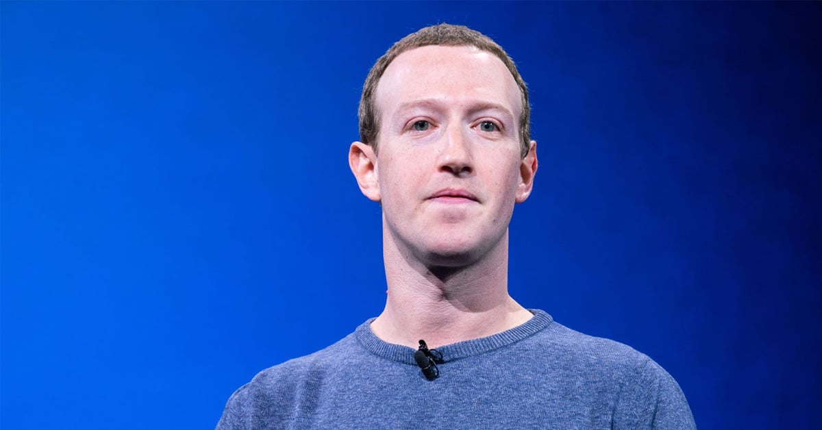 A person with short brown hair wearing a blue crewneck sweater stands in front of a plain blue background, looking slightly to the side. A small microphone is clipped to their collar.