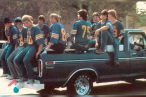 A group of boys wearing matching blue sports jerseys sit on the side and back of a pickup truck during the day. Some are sitting on the truck bed, while others sit along the edge. Trees and parked cars are visible in the background.