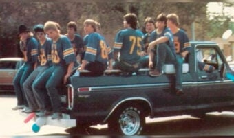 A group of boys wearing matching blue sports jerseys sit on the side and back of a pickup truck during the day. Some are sitting on the truck bed, while others sit along the edge. Trees and parked cars are visible in the background.