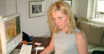 A woman with blonde hair and a gray sleeveless top sits at a desk, looking up at the camera. She is using a keyboard and mouse with a computer, and framed photos are visible on the wall behind her.