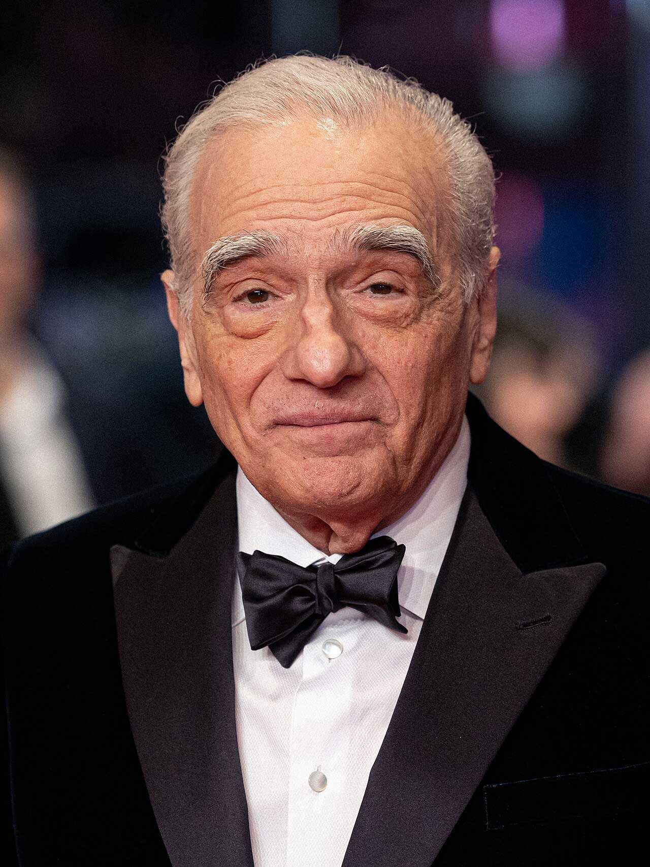 An elderly man with gray hair wearing a black tuxedo, white dress shirt, and black bow tie smiles slightly while standing in front of a blurred background at a formal event.