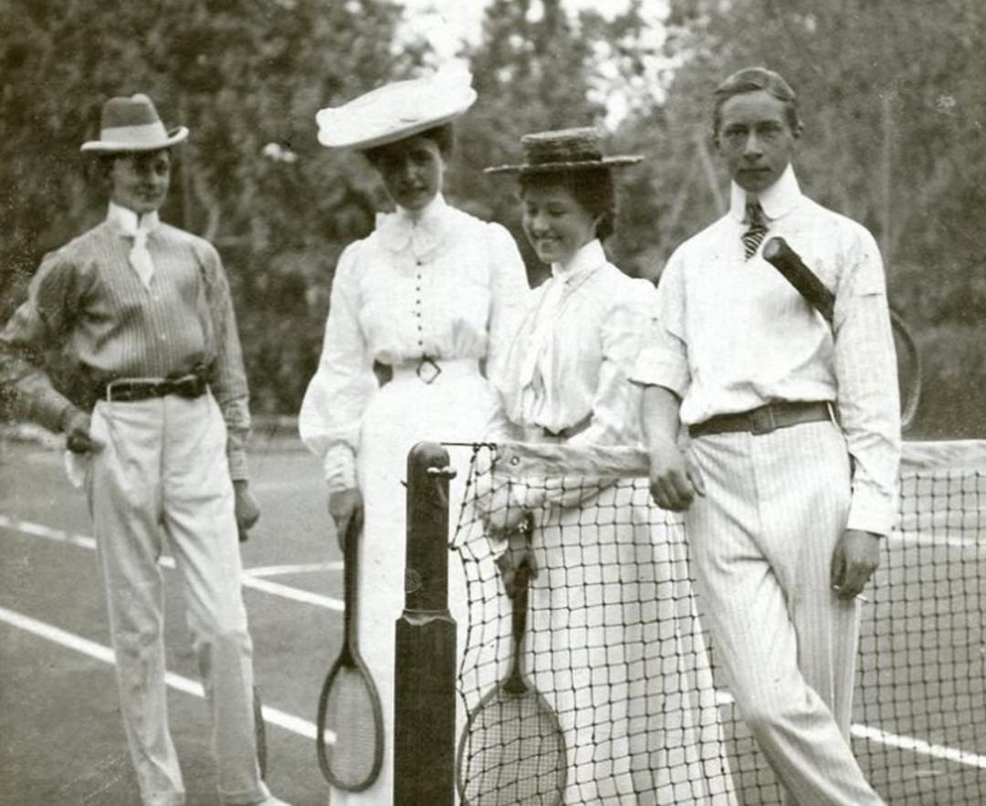 Four people in early 1900s attire pose with tennis rackets on a tennis court, two standing on each side of the net, all wearing hats and long-sleeved clothing. Trees are visible in the background.