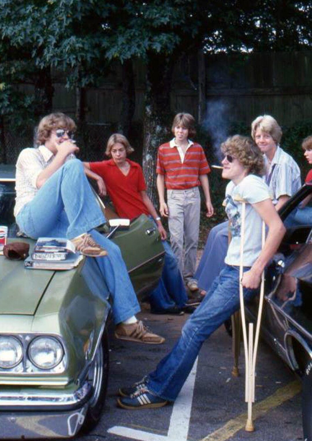 A group of teenage boys hang out by two parked cars. One sits on a car hood with magazines, another stands with crutches, and others stand nearby, all wearing casual 1970s-style clothes, chatting and relaxed. Trees and a fence are in the background.