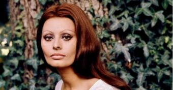 A woman with long, reddish-brown hair and striking eye makeup poses outdoors in front of a tree trunk and leafy green ivy, wearing a white top and gazing calmly at the camera.
