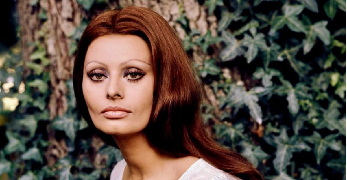 A woman with long, reddish-brown hair and striking eye makeup poses outdoors in front of a tree trunk and leafy green ivy, wearing a white top and gazing calmly at the camera.