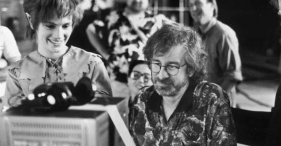 A group of people gather around a monitor, smiling and looking at the screen. The scene appears to be on a film set. One person with glasses and curly hair sits in front, while others stand behind, appearing cheerful and engaged.