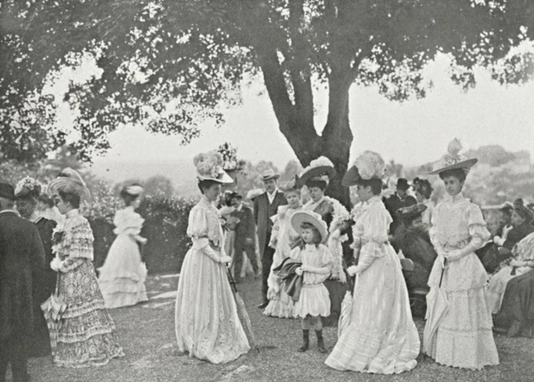 A black-and-white photo shows women and girls in elaborate Victorian-era dresses and hats gathered outdoors under a large tree, with men in suits and hats standing and seated in the background.