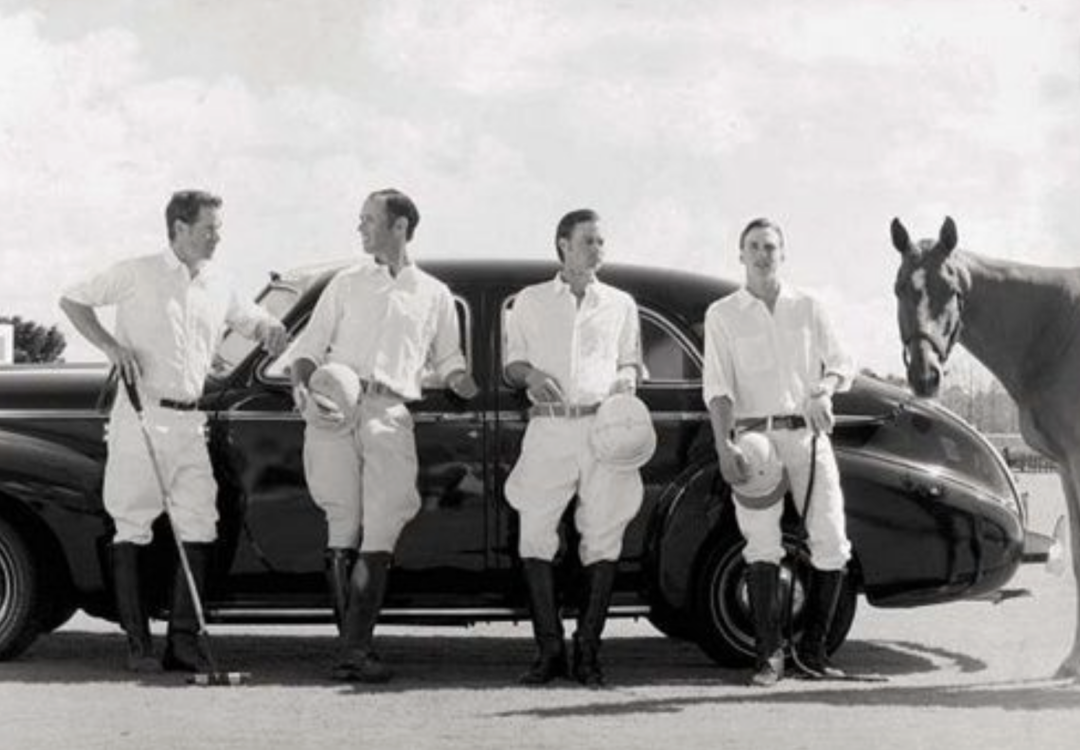 Four men in polo uniforms stand in front of a classic car, holding helmets and polo mallets. A horse stands on the right side of the image, near the group. The photo is in black and white.