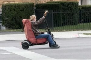 A person rides a scooter with a large, cushioned seat resembling an armchair on a city street, holding one arm up while passing by bushes and a brick building.