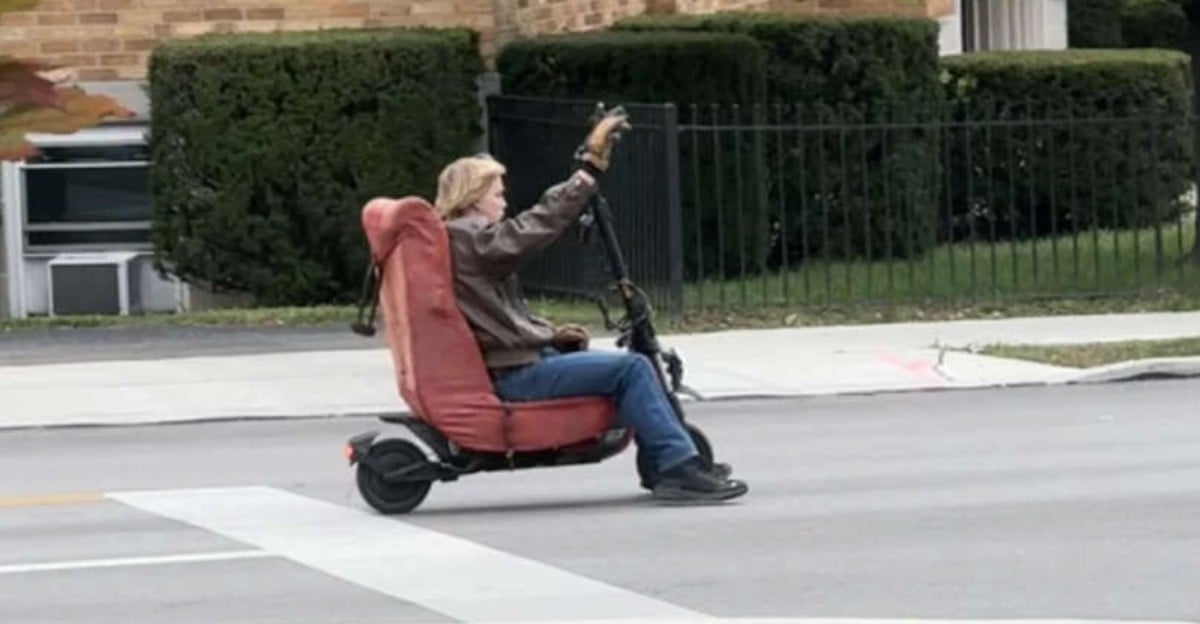 A person rides a scooter with a large, cushioned seat resembling an armchair on a city street, holding one arm up while passing by bushes and a brick building.