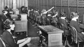 A row of women in vintage dresses sit at a large telephone switchboard, connecting calls with cords and plugs in a busy, early 20th-century office setting.