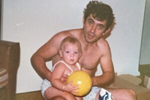 A shirtless man sits on the floor holding a small child with blonde hair who is holding a yellow ball. Toys and gifts are scattered around them, and the setting appears to be a cozy indoor room.