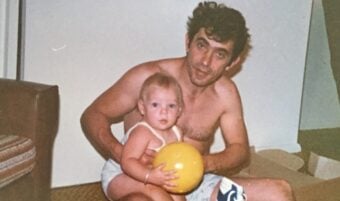 A shirtless man sits on the floor holding a small child with blonde hair who is holding a yellow ball. Toys and gifts are scattered around them, and the setting appears to be a cozy indoor room.