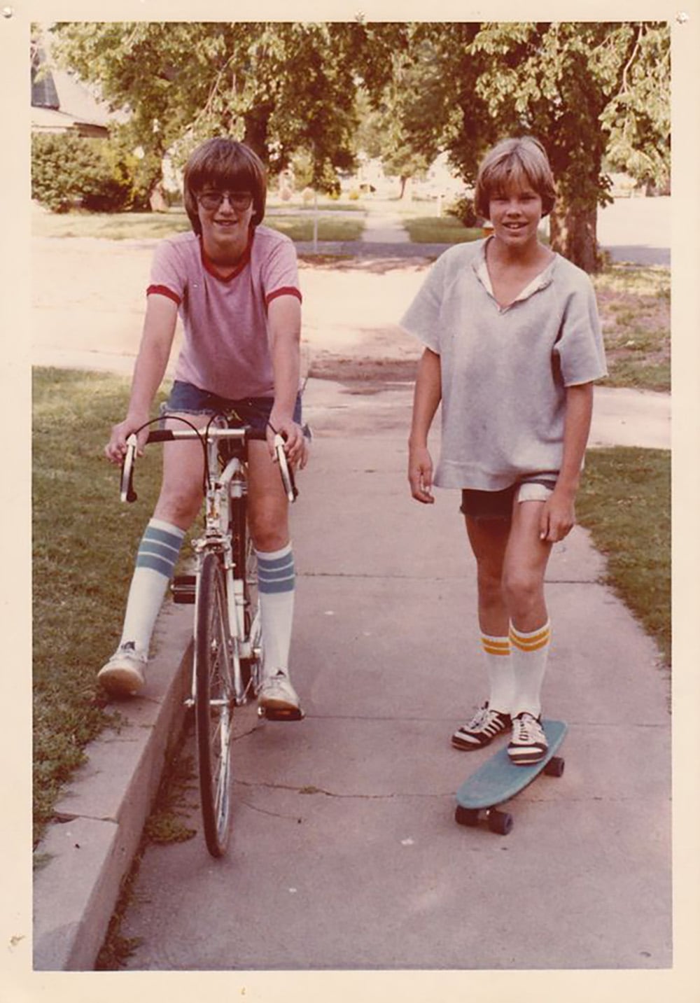 Two kids on a sidewalk, one riding a bicycle and the other standing on a skateboard. Both are wearing knee-high striped socks, shorts, and T-shirts. Trees and houses are visible in the background.