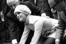A woman in a white outfit and hat presses her hands into wet cement while photographers, onlookers, and two men in suits kneeling beside her watch and take pictures.