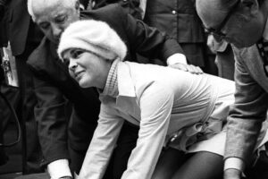 A woman in a white outfit and hat presses her hands into wet cement while photographers, onlookers, and two men in suits kneeling beside her watch and take pictures.