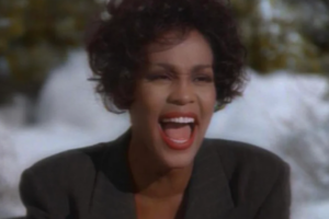 A woman with short curly hair and bright red lipstick sings passionately outdoors, wearing a dark blazer, with a blurred snowy background behind her.