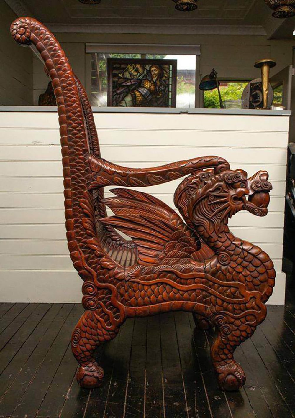 An intricately carved wooden chair shaped like a dragon, with detailed scales, wings, and a curled tail forming the chair's backrest and arms, set on a dark wooden floor in a well-lit room.
