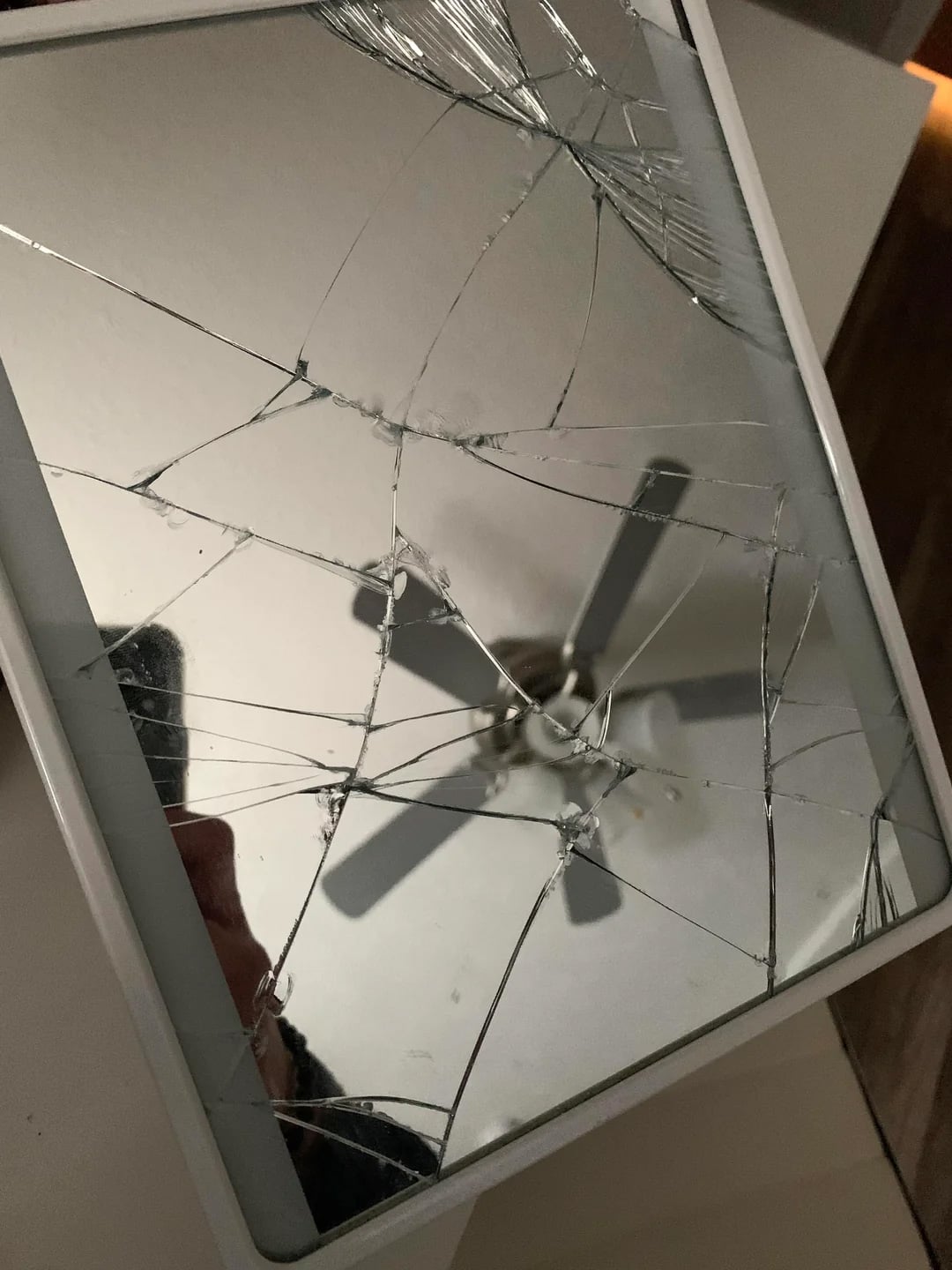 A tablet with a severely cracked screen reflects a ceiling fan and part of a person’s hand. The cracks radiate across the entire surface, creating a spiderweb pattern.