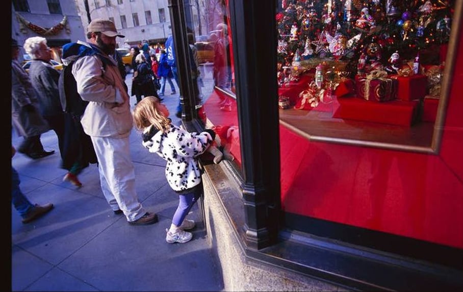 A young girl in a spotted coat gazes excitedly into a store window decorated with festive holiday ornaments and gifts. People walk by on the busy sidewalk behind her.