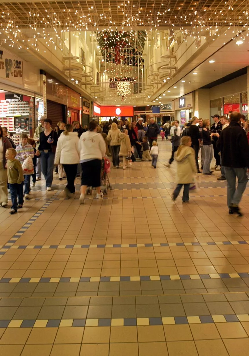 A busy shopping mall with people walking and shopping. Festive lights and decorations hang from the ceiling, and store fronts line both sides of the tiled walkway.