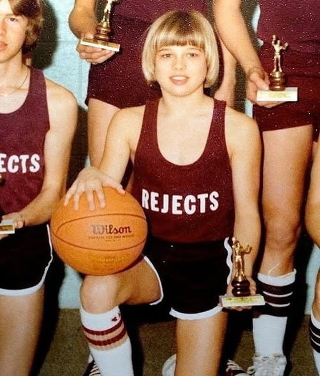 A young basketball player in a maroon "Rejects" jersey kneels on one knee, holding a basketball and a trophy, surrounded by teammates wearing matching uniforms and holding trophies.