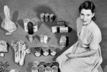 A woman in a striped dress sits on the floor, surrounded by neatly arranged groceries, including canned goods, boxed items, vegetables, and bags of flour or sugar, organized in rows on a carpeted surface.