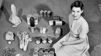 A woman in a striped dress sits on the floor, surrounded by neatly arranged groceries, including canned goods, boxed items, vegetables, and bags of flour or sugar, organized in rows on a carpeted surface.