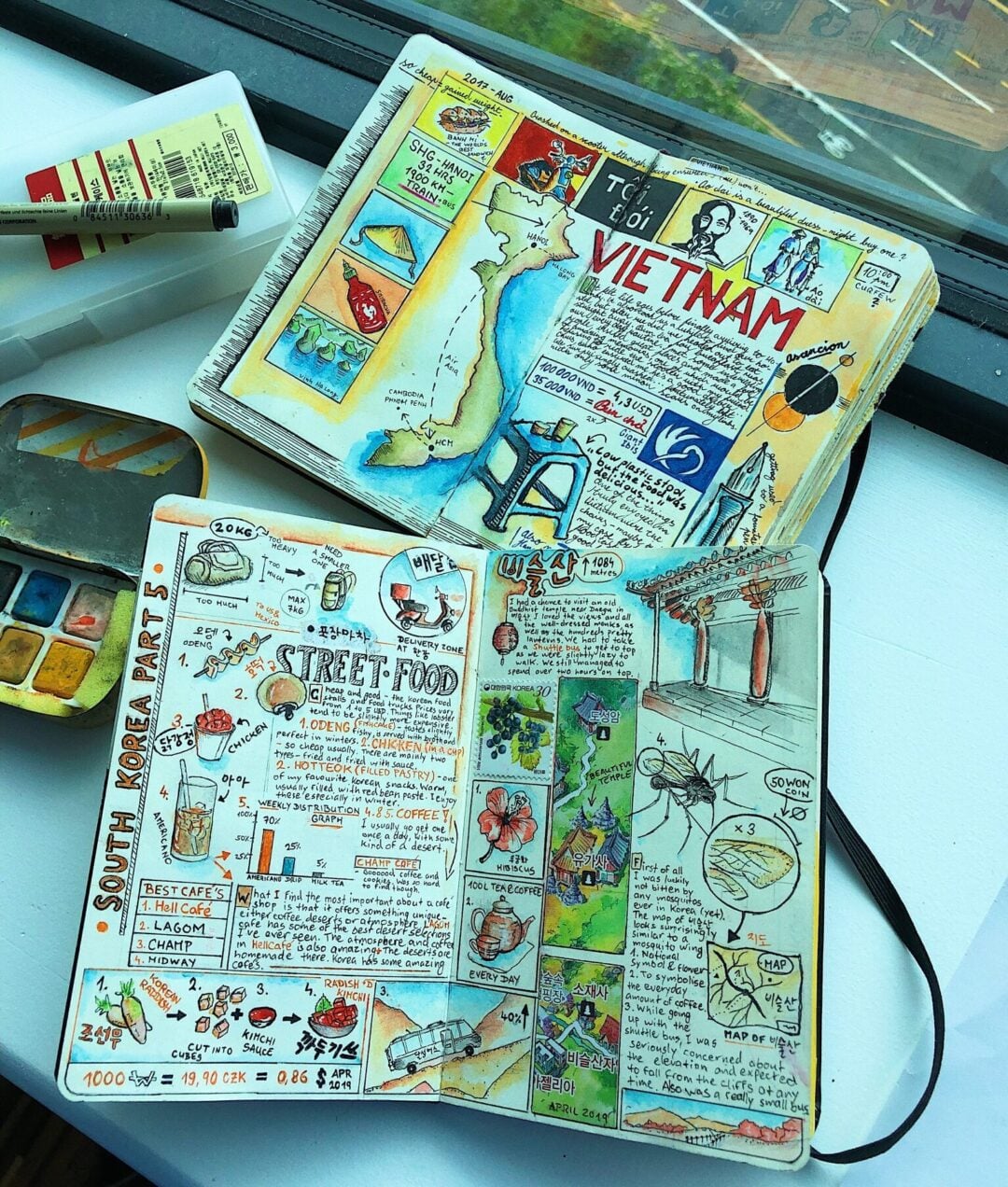 Two open travel journals filled with colorful sketches, maps, and notes about Vietnam and South Korea, featuring drawings of food, landmarks, and cultural icons. A watercolor set and paintbrush lies nearby on a white surface.