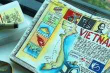 Two open travel journals filled with colorful sketches, maps, and notes about Vietnam and South Korea, featuring drawings of food, landmarks, and cultural icons. A watercolor set and paintbrush lies nearby on a white surface.