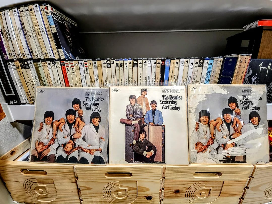Three vinyl records of The Beatles' "Yesterday and Today" album are displayed in front of shelves filled with tapes or cassettes. Each record shows a different version of the album cover.