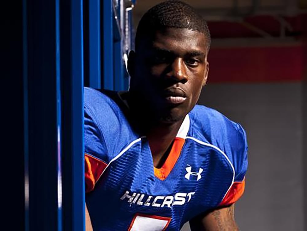 A football player wearing a blue "Hillcrest" jersey with orange and white accents stands in a locker room, looking seriously at the camera.